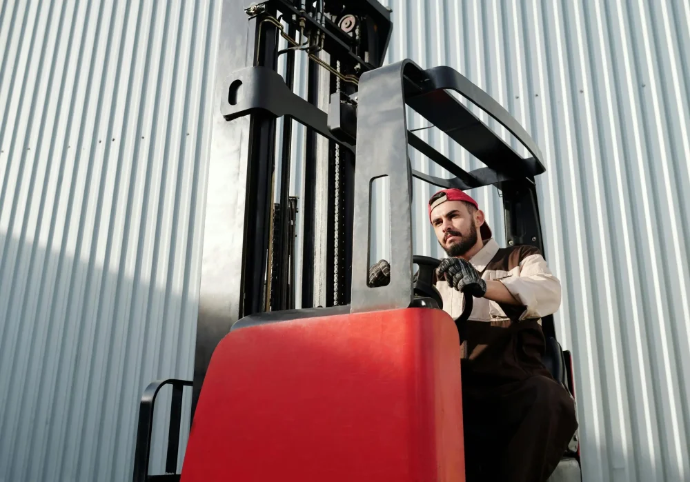 Forklift operator maneuvering a red stand-up forklift rental outside a metal building in Calgary, AB, working with Forklift Guy — forklift services calgary